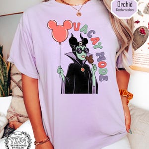 K&ouml;nnte beinhalten: Ein helllila T-Shirt mit einer Cartoon-Illustration einer Figur, die einen Mickey Mouse-Ballon und ein Eis h&auml;lt. Der Text "VACAY MODE" ist in bunten Buchstaben gehalten. Das Shirt hat auch den Text "Orchid Comfort colors" und "TDM Tees Designed by Megan".