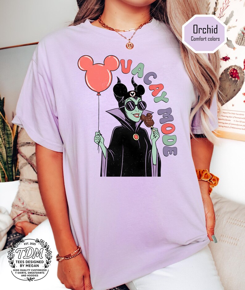 K&ouml;nnte beinhalten: Ein helllila T-Shirt mit einer Cartoon-Illustration einer Figur, die einen Mickey Mouse-Ballon und ein Eis h&auml;lt. Der Text "VACAY MODE" ist in bunten Buchstaben gehalten. Das Shirt hat auch den Text "Orchid Comfort colors" und "TDM Tees Designed by Megan".