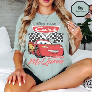 Puede incluir: Camiseta verde claro con un dise&ntilde;o de Rayo McQueen de Cars de Disney Pixar. El dise&ntilde;o incluye el logotipo de Cars, banderas a cuadros y el texto "Lightning McQueen". La camiseta tambi&eacute;n tiene el texto "Bay Comfort Colors".