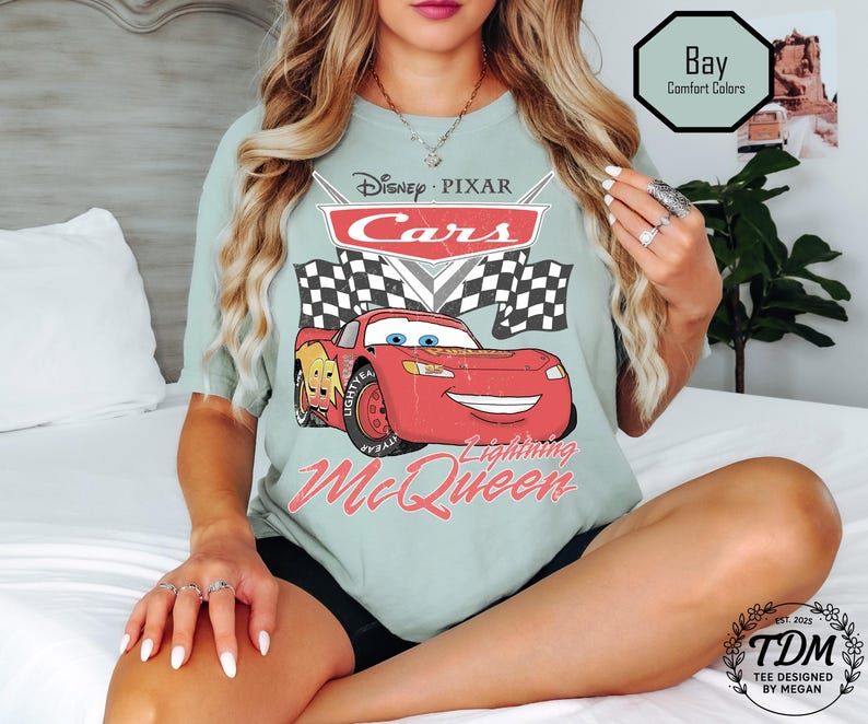 Puede incluir: Camiseta verde claro con un dise&ntilde;o de Rayo McQueen de Cars de Disney Pixar. El dise&ntilde;o incluye el logotipo de Cars, banderas a cuadros y el texto "Lightning McQueen". La camiseta tambi&eacute;n tiene el texto "Bay Comfort Colors".