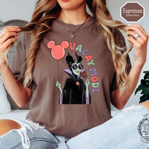 K&ouml;nnte beinhalten: Espressofarbenes T-Shirt mit einer Cartoon-Illustration eines Disney-B&ouml;sewichts, der einen Mickey-Mouse-Ballon und einen Eisriegel h&auml;lt. Der Text "VACAY MODE" ist um die Figur herum angeordnet. Das Shirt hat eine lockere Passform und geh&ouml;rt zur Comfort Colors-Kollektion.