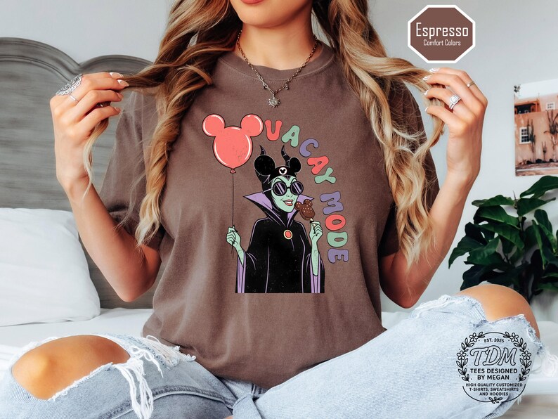 K&ouml;nnte beinhalten: Espressofarbenes T-Shirt mit einer Cartoon-Illustration eines Disney-B&ouml;sewichts, der einen Mickey-Mouse-Ballon und einen Eisriegel h&auml;lt. Der Text "VACAY MODE" ist um die Figur herum angeordnet. Das Shirt hat eine lockere Passform und geh&ouml;rt zur Comfort Colors-Kollektion.
