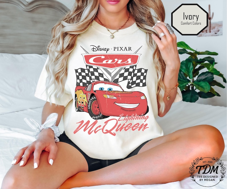 Puede incluir: Camiseta de color marfil con un dise&ntilde;o de Rayo McQueen de Cars de Disney Pixar. El dise&ntilde;o incluye el logotipo de Cars, banderas a cuadros y el texto "Lightning McQueen". La camiseta est&aacute; etiquetada como "Comfort Colors" y "Tee Designed by Megan".