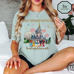 May include: A light green t-shirt with a colourful graphic of Cinderella's castle, the Epcot ball, and Disney characters. The text "WALT DISNEY WORLD" and "The most magical place on earth" are printed on the shirt. The shirt is labelled "Bay Comfort Colors".