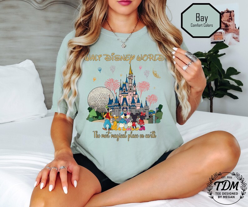 May include: A light green t-shirt with a colourful graphic of Cinderella's castle, the Epcot ball, and Disney characters. The text "WALT DISNEY WORLD" and "The most magical place on earth" are printed on the shirt. The shirt is labelled "Bay Comfort Colors".