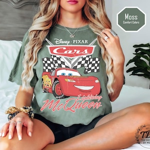 Puede incluir: Una camiseta verde musgo con un gr&aacute;fico de Rayo McQueen de Cars de Disney Pixar. El dise&ntilde;o incluye el logotipo de Cars, una bandera a cuadros y el texto "Lightning McQueen". La camiseta tambi&eacute;n tiene el texto "Moss Comfort Colors".
