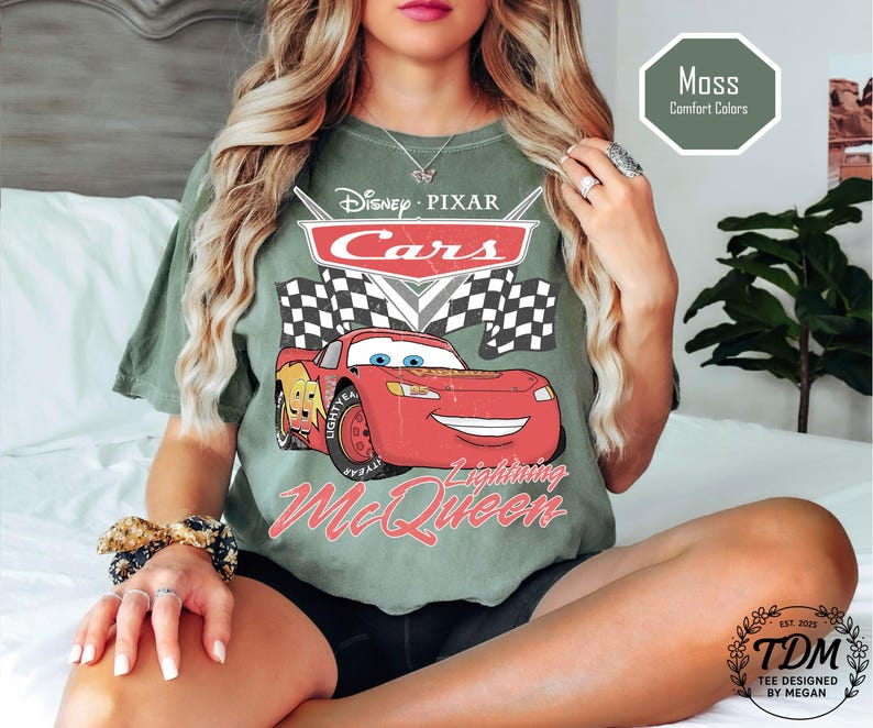 Puede incluir: Una camiseta verde musgo con un gr&aacute;fico de Rayo McQueen de Cars de Disney Pixar. El dise&ntilde;o incluye el logotipo de Cars, una bandera a cuadros y el texto "Lightning McQueen". La camiseta tambi&eacute;n tiene el texto "Moss Comfort Colors".