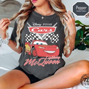 Puede incluir: Camiseta gris oscuro con un gr&aacute;fico de Rayo McQueen de Cars de Disney Pixar. El dise&ntilde;o incluye el logotipo de Cars, una bandera a cuadros y el texto "Lightning McQueen". La camiseta tambi&eacute;n tiene el texto "Pepper Comfort Colors".