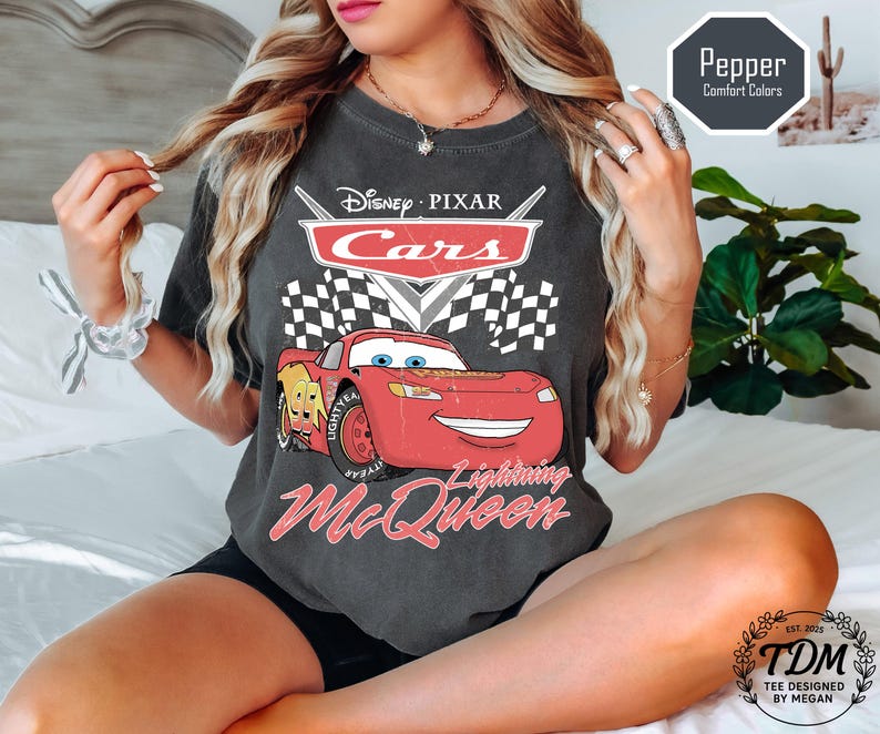 Puede incluir: Camiseta gris oscuro con un gr&aacute;fico de Rayo McQueen de Cars de Disney Pixar. El dise&ntilde;o incluye el logotipo de Cars, una bandera a cuadros y el texto "Lightning McQueen". La camiseta tambi&eacute;n tiene el texto "Pepper Comfort Colors".