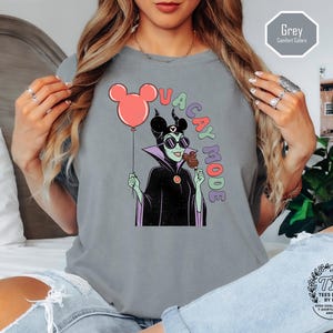K&ouml;nnte beinhalten: Graues T-Shirt mit einer Cartoon-Illustration einer Figur mit Sonnenbrille, die einen roten Mickey-Mouse-Ballon und einen Schokoladen-Snack h&auml;lt. Die Worte "VACAY MODE" sind in bunten Buchstaben auf das Shirt gedruckt. Das Shirt hat einen Rundhalsausschnitt.