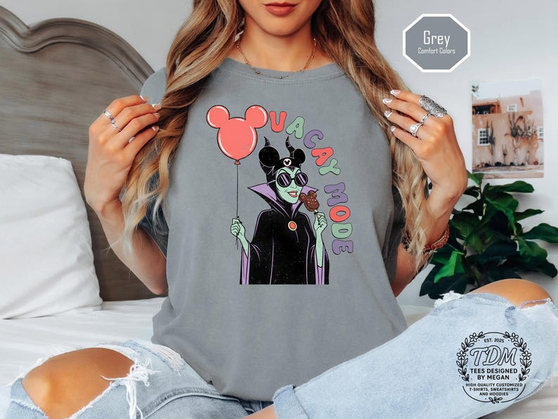 K&ouml;nnte beinhalten: Graues T-Shirt mit einer Cartoon-Illustration einer Figur mit Sonnenbrille, die einen roten Mickey-Mouse-Ballon und einen Schokoladen-Snack h&auml;lt. Die Worte "VACAY MODE" sind in bunten Buchstaben auf das Shirt gedruckt. Das Shirt hat einen Rundhalsausschnitt.