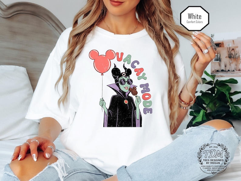 K&ouml;nnte beinhalten: Wei&szlig;es T-Shirt mit einer Grafik eines Zeichentrick-B&ouml;sewichts, der einen Mickey-Mouse-Ballon und ein Eis h&auml;lt. Der Text "VACAY MODE" ist um die Figur herum angeordnet. Das Shirt hat eine lockere Passform und ist mit "Comfort Colors" gekennzeichnet.