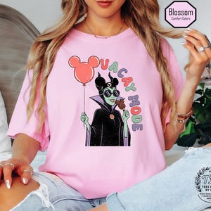 K&ouml;nnte beinhalten: Hellrosa T-Shirt mit einer Cartoon-Illustration einer Figur mit Sonnenbrille, die einen Mickey Mouse-Ballon und ein Eis h&auml;lt. Der Text "VACAY MODE" ist &uuml;ber der Figur angeordnet. Das T-Shirt hat eine lockere Passform.