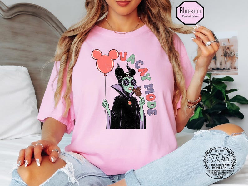 K&ouml;nnte beinhalten: Hellrosa T-Shirt mit einer Cartoon-Illustration einer Figur mit Sonnenbrille, die einen Mickey Mouse-Ballon und ein Eis h&auml;lt. Der Text "VACAY MODE" ist &uuml;ber der Figur angeordnet. Das T-Shirt hat eine lockere Passform.
