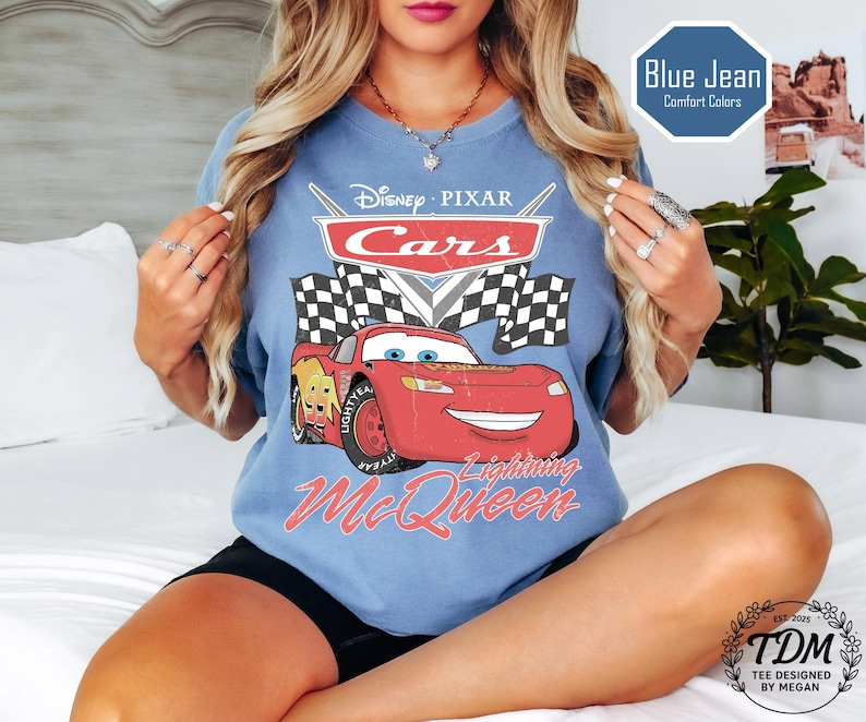 Puede incluir: Camiseta azul claro con el logotipo de Disney Pixar Cars y Lightning McQueen. El dise&ntilde;o incluye banderas a cuadros y el texto "Lightning McQueen". La camiseta tambi&eacute;n tiene el texto "Blue Jean Comfort Colors".