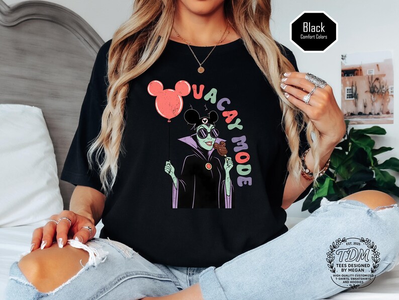 K&ouml;nnte beinhalten: Schwarzes T-Shirt mit einem Cartoon-Motiv einer gr&uuml;n geh&auml;uteten Figur mit Sonnenbrille, die ein Eis und einen Mickey-Mouse-Ballon h&auml;lt. Der Text "VACAY MODE" ist in bunten Buchstaben gehalten. Das Shirt ist mit "Comfort Colors" gekennzeichnet.
