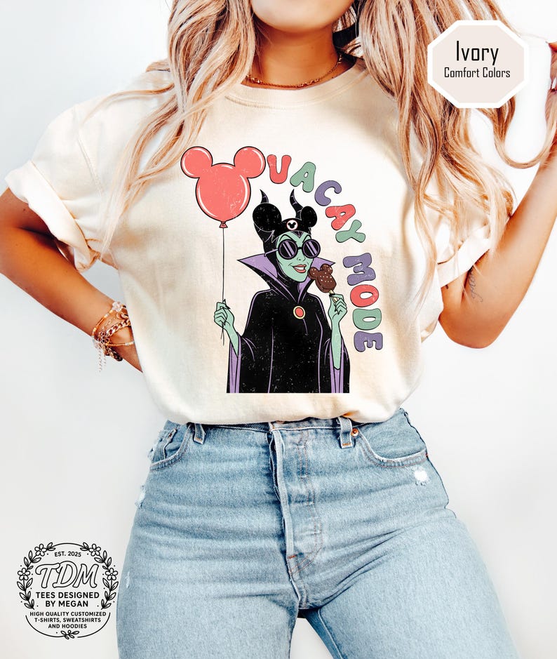 K&ouml;nnte beinhalten: Cremefarbenes T-Shirt mit einer Cartoon-Illustration einer Figur, die einen Mickey-Mouse-Ballon und einen Schokoladen-Snack h&auml;lt. Der Text "VACAY MODE" steht &uuml;ber der Figur. Das Shirt hat einen Rundhalsausschnitt und kurze &Auml;rmel.