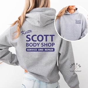 Puede incluir: Una sudadera con capucha gris jaspeado con un bolsillo delantero y una capucha con cordón. La parte trasera de la sudadera con capucha presenta el texto "Keith SCOTT BODY SHOP SERVICE AND REPAIR" en azul marino. Una versión más pequeña del logotipo está en la parte delantera.