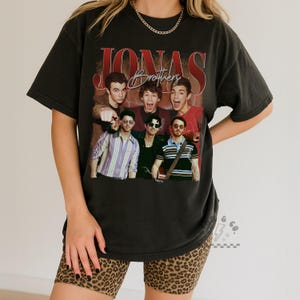 May include: A black short-sleeve t-shirt featuring a large graphic print of the Jonas Brothers. The design includes the text JONAS in large red letters and Brothers in script, with multiple photographic portraits of the band members below.