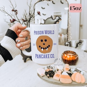 Puede incluir: Taza blanca de 15 oz con el texto "PRISON WORLD PANCAKE CO." y un gráfico de panqueque sonriente. Una galleta gris en forma de murciélago descansa en el borde. La taza es parte de una colección temática de Halloween.