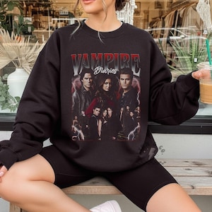May include: A black crewneck sweatshirt featuring a graphic print of the television show Vampire Diaries. The design includes a collage of characters from the series, red and white text, and lightning bolt accents on a dark background.
