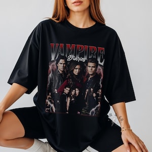 May include: A black oversized t-shirt featuring a graphic print of the cast from The Vampire Diaries. The text VAMPIRE DIARIES is printed in a bold red and white gradient font above the characters, set against a dark, moody background.
