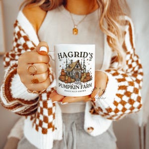 May include: White ceramic mug featuring the text "Hagrid's Pumpkin Farm" with a graphic of a house and pumpkins. The mug has a handle and is held by a person wearing a brown and white checkered cardigan.