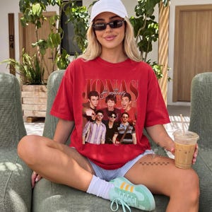 May include: A red short-sleeve t-shirt featuring a graphic print of the Jonas Brothers with the text JONAS BROTHERS in a bold serif font. The shirt has a crew neckline and a relaxed fit, showcasing a vintage-style collage of the band members.