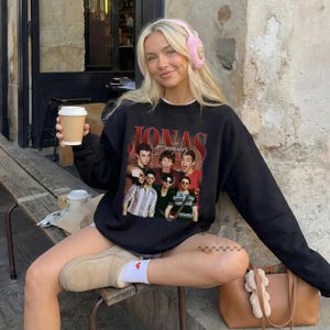 May include: A black long-sleeve sweatshirt featuring a graphic print of the Jonas Brothers with red text above the image. The sweatshirt is worn with light tan slip-on shoes, white socks with a small red heart, and pink knit headphones.