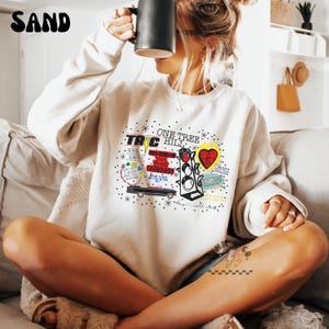 May include: Sand-colored sweatshirt featuring a graphic design with the text "One Tree Hill." The design includes a traffic light, a heart, and other elements. The sweatshirt is a neutral color and has a crew neck.