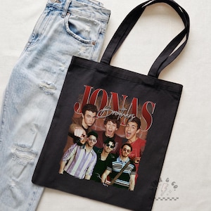 May include: A black tote bag featuring a graphic print of the Jonas Brothers with the text "JONAS Brothers" in red and white lettering. The bag has two black shoulder straps and is displayed next to a pair of light blue distressed denim jeans.