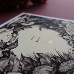 May include: A black and white floral illustration with gold foil lettering that reads "Live Slow Die Old".