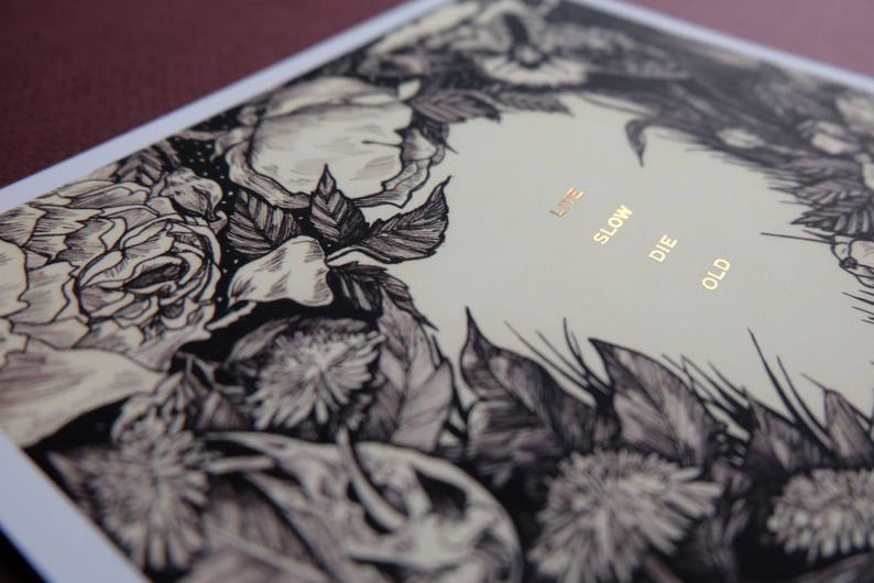 May include: A black and white illustration of flowers with the text "Live Slow Die Old" printed in gold on a cream background.