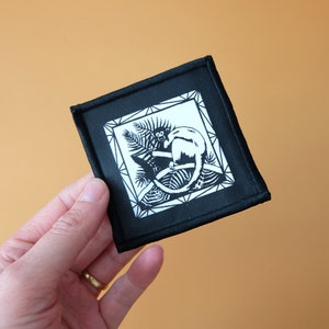 May include: Black and white embroidered patch featuring a monkey sitting in a tree. The patch is square and has a black border.
