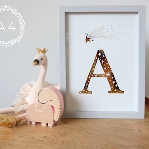 May include: A framed print with a gold foil letter A decorated with stars and a shooting star. The print is on a white background and is framed in a light gray frame. The print is sitting on a wooden table with a pink stuffed swan and a wooden elephant.
