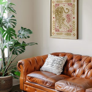 May include: A brown leather sofa with a white patterned pillow. A framed print of a tiger with gold and red details hangs on the wall above the sofa. A large green plant is in a woven basket on the floor next to the sofa.