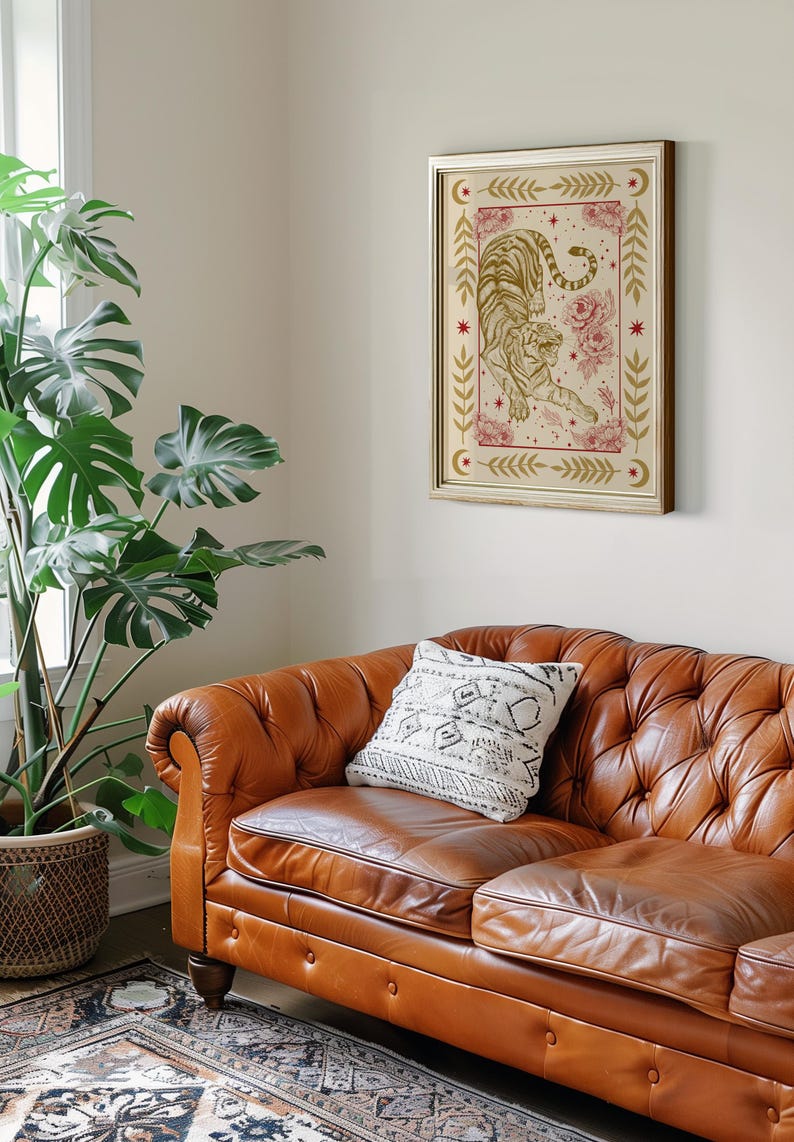 May include: A brown leather sofa with a white patterned pillow. A framed print of a tiger with gold and red details hangs on the wall above the sofa. A large green plant is in a woven basket on the floor next to the sofa.