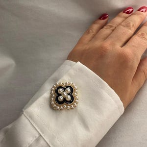 May include: A gold-toned cufflink with a black clover design, surrounded by small pearls. The cufflink is attached to a white dress shirt cuff. The person's hand with red nail polish is visible.