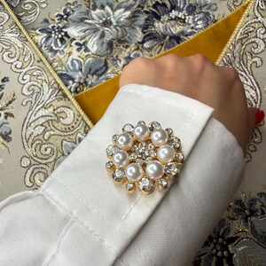 May include: A circular gold-tone cufflink featuring a central cluster of clear crystals surrounded by a ring of eight white faux pearls, with additional small clear crystals set along the outer scalloped edge, attached to a white fabric shirt cuff.