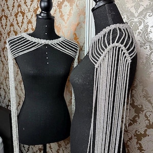 May include: A silver shoulder wrap displayed on a black mannequin. The wrap features a beaded collar and long, dangling strands. The mannequin is set against a patterned wallpaper background, with a second mannequin visible in a mirror.