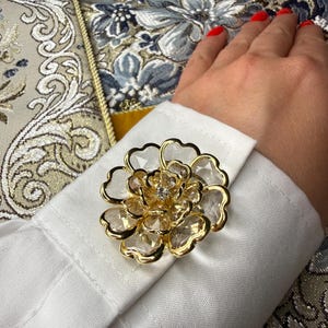 May include: A gold-toned cufflink featuring a multi-layered flower design with clear, faceted crystal petals and a central clear stone. The accessory is attached to the white fabric cuff of a long-sleeved shirt, showcasing a detailed floral shape.