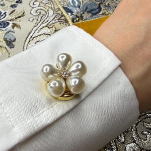 May include: A gold-toned cufflink featuring a flower design with five teardrop-shaped faux pearls arranged around a central cluster of small clear crystals, attached to the white fabric cuff of a long-sleeved shirt.