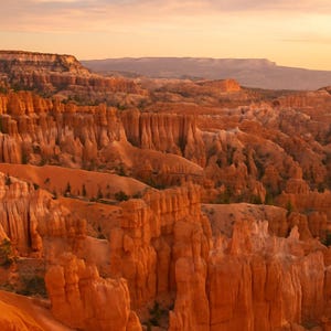 May include: A scenic landscape of Bryce Canyon National Park at sunset. The image showcases towering, orange-hued rock formations, with a warm, golden light illuminating the scene. The sky transitions from orange to a soft, pale yellow.
