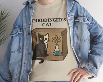 May include: Beige t-shirt with the text "Schrödinger's Cat" above a cartoon illustration of a cat, an atom symbol, and a flask. The cat is half black and half white, sitting inside a cardboard box. The person is wearing a blue denim jacket.