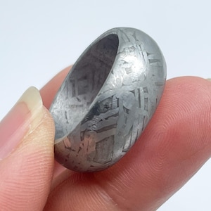 May include: A close-up of a silver ring with a unique patterned design. The ring is held between two fingers against a white background. The ring's surface has a textured appearance.