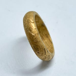 Peut inclure: Une bague dorée avec une surface texturée. La bague a un design de type alliance et semble être en métal. La bague est présentée sur une surface blanche.
