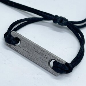 May include: A close-up of a bracelet featuring a rectangular, textured, gray metal bar with two holes, connected to a black cord. The cord is knotted on both sides of the bar, forming the bracelet's adjustable design.