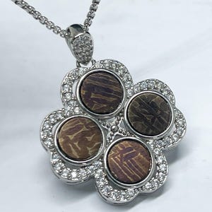 Puede incluir: Un collar con colgante plateado con un diseño de trébol de cuatro hojas. Cada hoja es una piedra circular con un patrón marrón y morado, rodeada por un halo de cristales transparentes. La cadena del collar también es plateada.