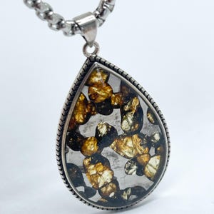 Puede incluir: Un collar con colgante en forma de lágrima con una cadena plateada. El colgante presenta un recinto transparente, similar al vidrio, lleno de piedras irregulares, de color marrón dorado y negro. El colgante está enmarcado con un borde plateado en forma de cuerda.