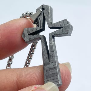 以下が含まれることがあります： 星型の切り抜きがある、質感のあるグレーの金属製の十字架ペンダント。ペンダントはシルバーチェーンに吊り下げられています。十字架は、白い背景に対して人の指で保持されています。
